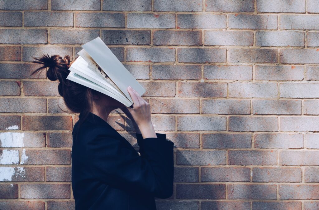 perfil de uma mulher em frente a uma parede de tijolinho segurando um livro na frente do rosto