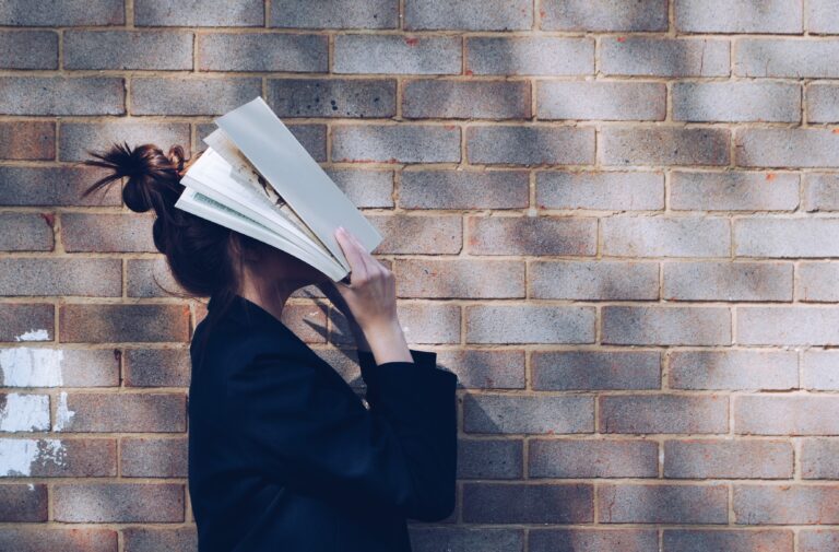 perfil de uma mulher em frente a uma parede de tijolinho segurando um livro na frente do rosto