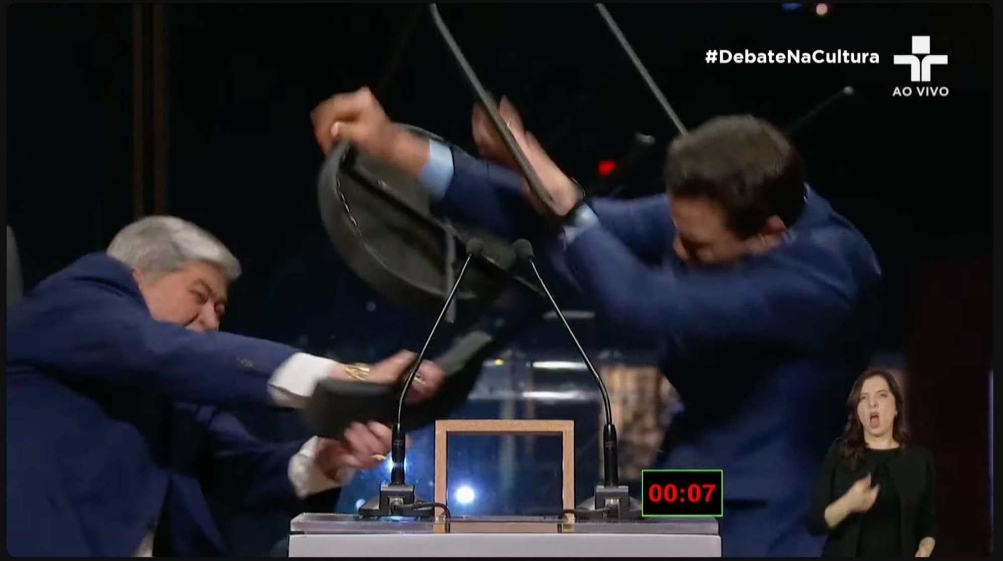 Este é um frame do video do debate entre candidatos a prefeito de SP. À esquerda temos Datena, de terno azul, consumando uma cadeirada em Pablo Marçal, à direita, que está com os braços levantados, em defesa. A parte da direita do frame está com um pouco de blur por causa do movimento. No frame é possível ver que as pernas da cadeira quebraram no braço de Marçal, que estava respondendo Datena. O timestamp mostra que Marçal ainda tinha sete segundos de resposta, que foi interrompida com a cadeirada. A intérprete de Libras está de boca aberta, não sei se por causa da tradução ou por causa do evento em tela.