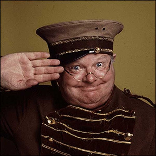 Imagem do apresentador Benny Hill. Ele está de óculos, usando um quepe e um uniforme de guarda.